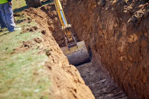 Koparka pogłębia wykop, sięgając do strefy przemarzania gruntu. Pracownicy nadzorują prace.