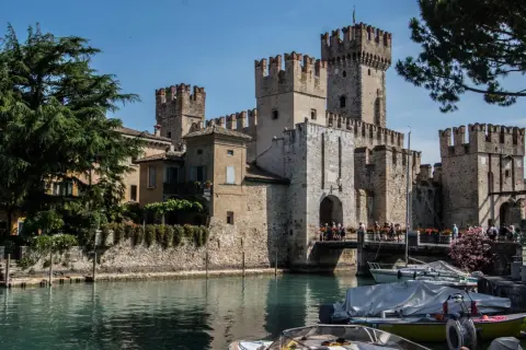 Il castello di Moniga si affaccia sull'acqua, con torri merlate e un ponte pedonale affollato. Barche ormeggiate completano la scena.