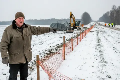 Mężczyzna wskazuje na teren budowy drogi, zastanawiając się, czy gmina może zabrać część działki. W tle koparka i pracownicy.