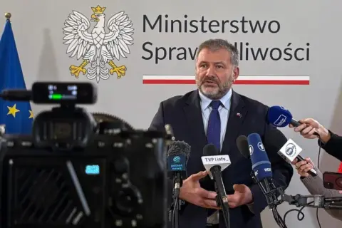 Minister sprawiedliwości przemawia do mediów, otoczony flagami Polski i UE. Dziennikarze zbierają materiał do reportażu o wyroku TSUE.
