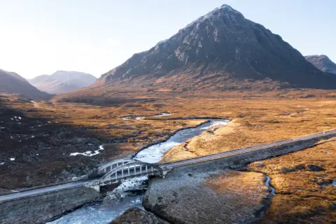 Majestätische Berge der Schottland Highlands, ein Fluss schlängelt sich durch das goldene Gras, überquert von einer Brücke.
