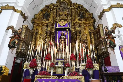 Un altar barroco con imágenes religiosas, velas y flores rojas. Destaca una figura central de Cristo, que parece ser un quinario, rodeado de figuras de la Virgen y otros santos.