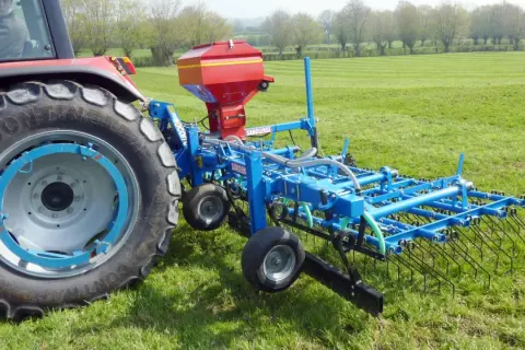 Tracteur rouge avec semoir bleu pour le sursemis de prairie. L'outil prépare le sol avant de semer de nouvelles herbes.