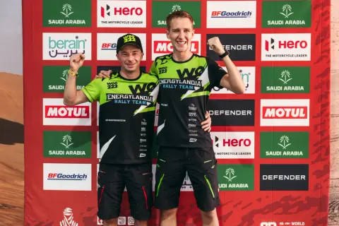 Dwóch zawodników Energylandia Rally Team pozuje z podniesionymi pięściami na tle baneru z logo sponsorów.