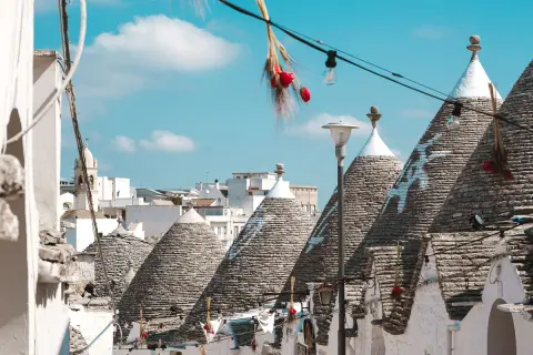 Scorci di Alberobello con i caratteristici trulli, alcuni decorati con simboli bianchi, sotto un cielo azzurro. Una scalinata cuori conduce tra le abitazioni uniche.