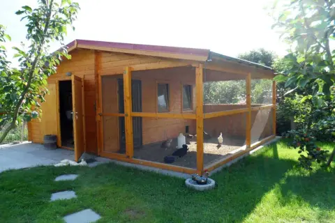 Holz-Gartenhaus mit integriertem Hühnerstall und Freigehege. Hühner picken im Sand, ein Hahn sitzt im Wasserbad.