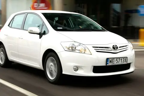 Biała Toyota Auris w ruchu miejskim, z widocznym kierowcą za kierownicą.