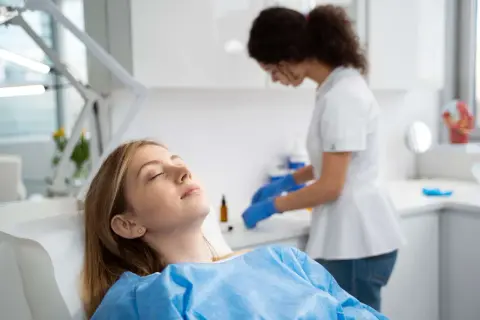 Paziente sdraiata con gli occhi chiusi, forse con un leggero gonfiore guancia dopo anestesia dentista, mentre un'assistente prepara strumenti.