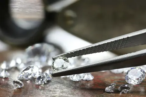 Szczypce trzymają błyszczący diament, obok leżą inne kamienie. To nie jest kaboszon, lecz brylant.