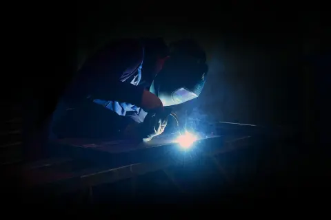 Spawacz w masce ochronnej używa elektrody do żeliwa, tworząc jasny łuk świetlny i dym.