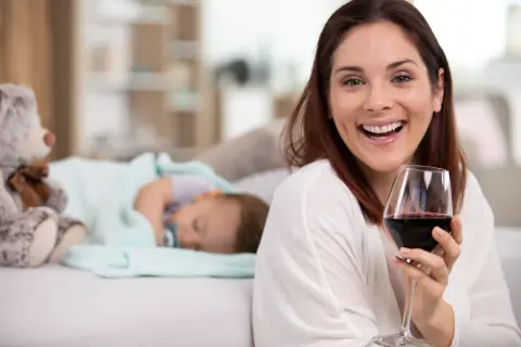 Uśmiechnięta kobieta z kieliszkiem czerwonego wina, podczas gdy dziecko śpi obok. Temat: karmienie piersią a alkohol.