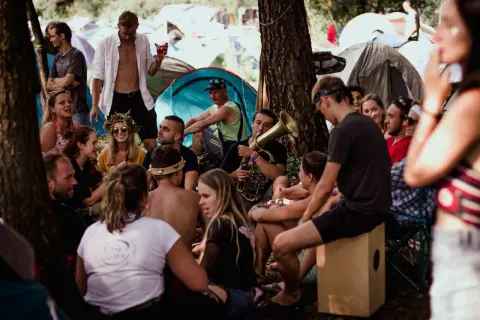 Grupa ludzi relaksuje się na festiwalu Pannonica, jeden z nich gra na puzonie. W tle widać namioty.