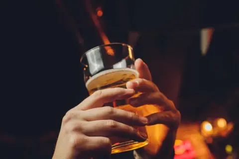 Dwie dłonie trzymające szklankę piwa, w tle rozmyte światła i chorągiewki. Czy piwo tuczy?