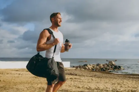 Uśmiechnięty mężczyzna z torbą sportową i bidonem na plaży. Zastanawia się, glutamina co to, czy pomoże mu w regeneracji po treningu.
