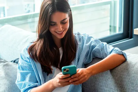 Une jeune femme souriante lit un livre à lire sur téléphone, confortablement installée sur un canapé gris.