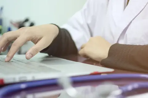 Lekarz wpisuje dane pacjenta na laptopie, co oznacza skierowanie na cito. Stetoskop leży obok.