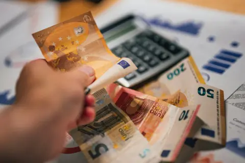 Dłoń trzyma banknot 50 euro nad stosem innych banknotów i kalkulatorem. To symbol prowizji i finansów.
