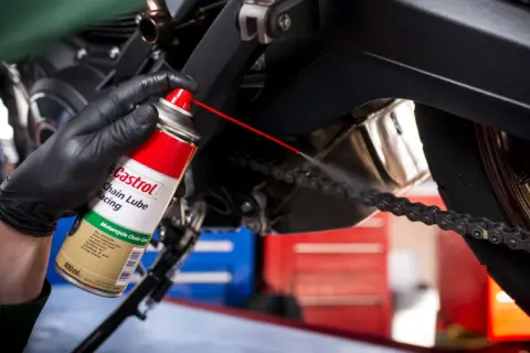 Ręka w czarnej rękawiczce rozpyla smar Castrol na łańcuch motocykla.