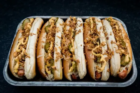 Sechs Hot Dogs in einem Blech, belegt mit Gurken, Sauerkraut, Senf und knusprigen Zwiebeln – die perfekten Hot Dog Zutaten für jeden Geschmack.