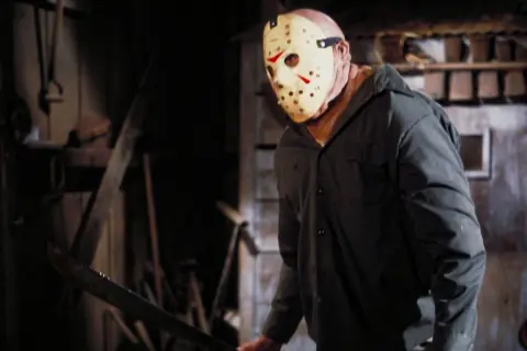 Postać w masce hokejowej, z maczetą w ręku, groźnie wygląda w mroku. Klasyczny slasher horror.