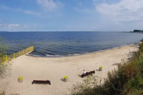 Plaża dla psów z łagodnym zejściem do morza. Na piasku ławki i kosze. W oddali widać falochron.