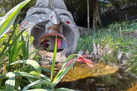 Scultura di una testa gigante con bocca spalancata nel giardino botanico Gardone Riviera, con piante rigogliose e uno stagno.