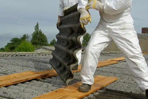 Arbeiter in Schutzanzügen entfernen eine Wellplatte. Bis in die 1990er Jahre wurde Asbest verbaut.
