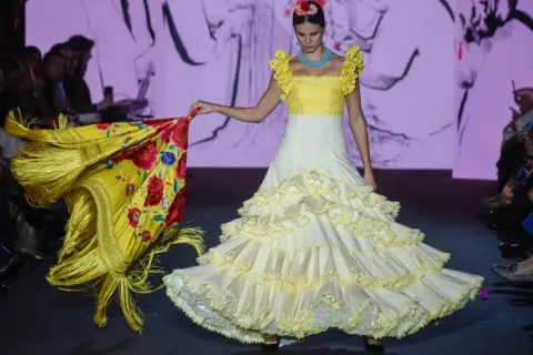 Modelo desfila en la pasarela We Love Flamenco con un vestido blanco de lunares amarillos y volantes, portando un mantón amarillo con flores rojas.