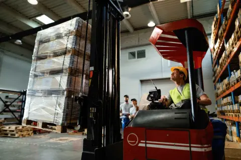 Operator wózka widłowego przygotowuje transport drobnicowy. Na paletach leżą paczki owinięte folią.