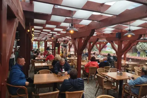 Zadaszony ogródek restauracyjny z drewnianymi belkami, lampami i siedzącymi przy stołach ludźmi.
