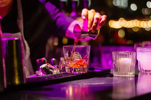 Barman nalewa bursztynowy cordial drink do szklanki z lodem. W tle kolorowe światła.