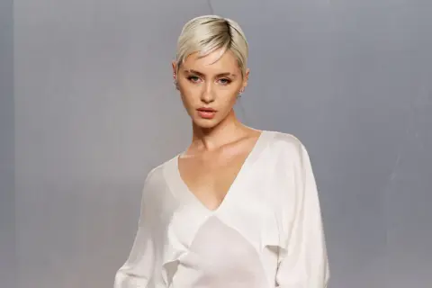 Iris Law, con su corte de pelo rubio platino y un elegante vestido blanco, posa con una mirada cautivadora.