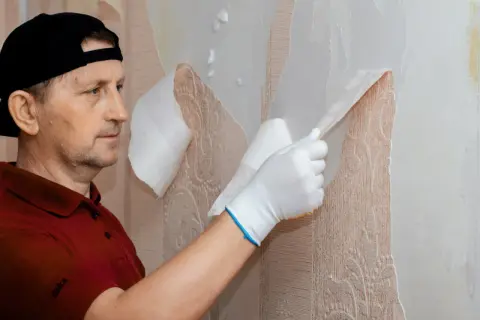 Mężczyzna w czapce i rękawiczkach zrywa starą tapetę, odsłaniając wzorzystą powierzchnię.