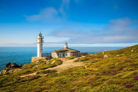 Faro blanco y edificio antiguo en Cabo Touriñán, con mar azul y cielo nublado.