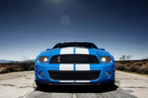 Niebieski Shelby GT500 z białymi pasami na masce i dachu, zaparkowany na pustynnej drodze pod błękitnym niebem.