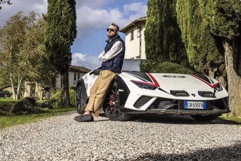 Mężczyzna opiera się o białe Lamborghini Sterrato z czerwono-czarnymi akcentami, zaparkowane na żwirowej drodze wśród drzew.