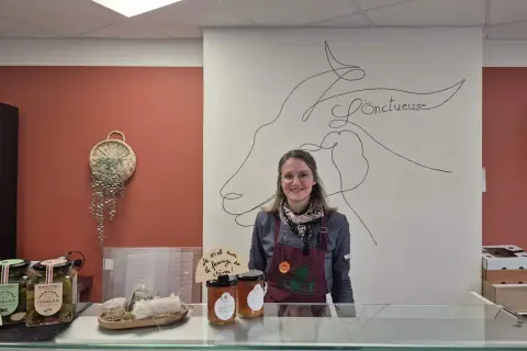 Une vendeuse souriante derrière un comptoir, avec des cornichons, du miel et du fromage de chèvre, peut-être du fromage Saint-Aubin, en vente.