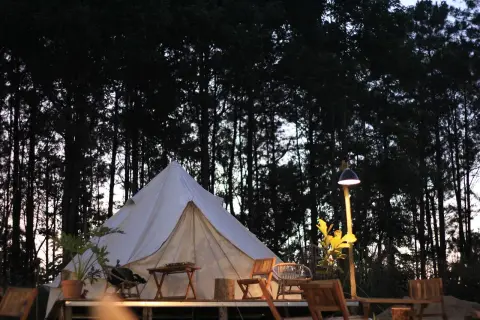 En vit glampingtält står uppsatt bland höga träd i skymningen. Utanför tältet finns sittmöbler och ett litet bord, redo för avkoppling. En lampa lyser upp området.