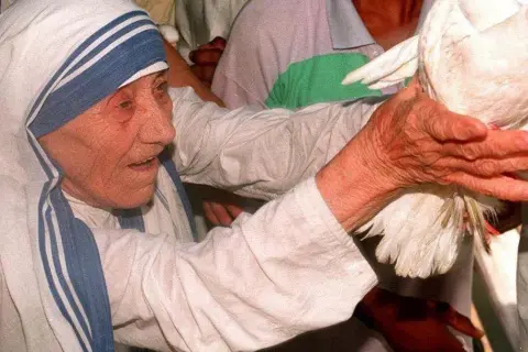Matka Teresa z Kalkuty, otoczona ludźmi, trzyma gołębia. Wokół niej rozchodzą się plotki i zarzuty, ale ona wciąż emanuje spokojem.