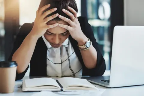 Kvinna med händerna i håret, blicken sänkt över en öppen bok. Känslan av stress på jobbet är påtaglig. En kaffekopp och laptop finns i närheten.