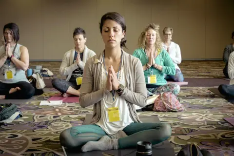 Frauen in einer Gruppe praktizieren transzentale Meditation, sitzen im Schneidersitz mit gefalteten Händen und geschlossenen Augen.