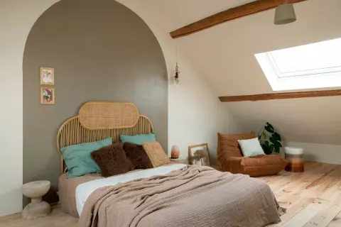 Gezellige slaapkamer verbouwen met rieten hoofdbord, zachte plaids en een comfortabele fauteuil onder het dakraam.