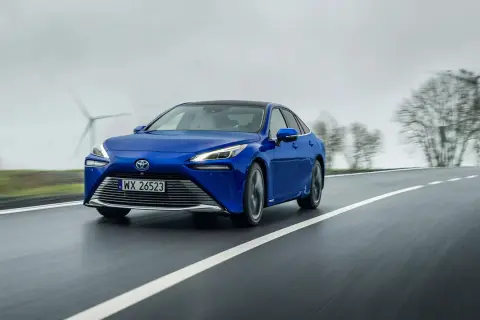 Niebieski sedan z napędem wodorowym jedzie po mokrej drodze, w tle widać wiatraki i drzewa.