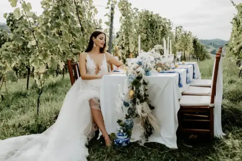 Braut in weißem Kleid sitzt am Tisch im Weinberg. Eine romantische Kulisse für eine weingut hochzeit.