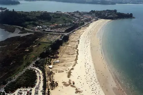 Vista aérea de una playa grande y concurrida, con arena dorada, mar azul y un pueblo costero al fondo.