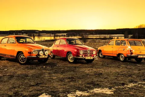 Tre klassiska Saab-bilar, en röd och två orange, står uppradade i solnedgången. En laban bil som väcker nostalgi.