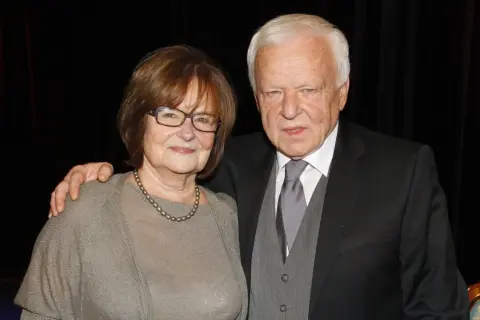 Anna Opani w okularach i z perłami na szyi, obok elegancko ubranego mężczyzny w garniturze.