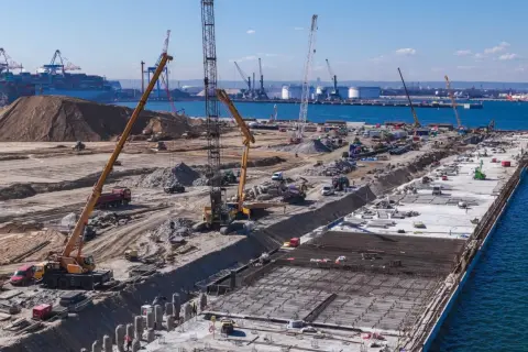 Budowa dużego portu w Japonii. Dźwigi, koparki i ciężarówki pracują na placu budowy, tworząc nową infrastrukturę.