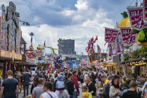Buntes Volksfest mit vielen Menschen, Fahrgeschäften und Buden. Ein Hamburger-Stand ist nicht zu sehen, aber die Stimmung ist ausgelassen.