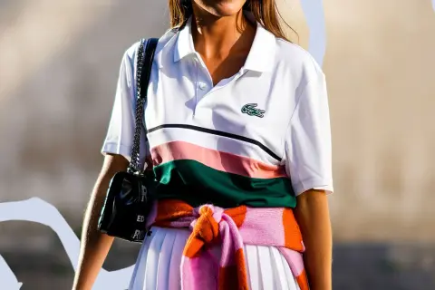 Frau trägt weißes Lacoste Poloshirt mit Streifen, schwarze Tasche und bunte Strickjacke um die Taille gebunden.
